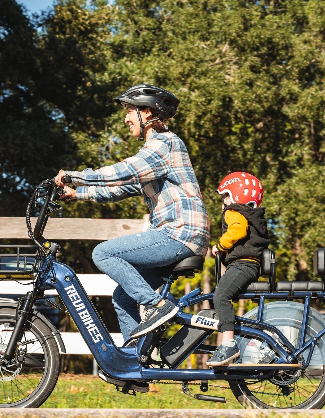 Person riding a revi flux blue cargo electric bike with a child in a seat, surrounded by greenery.