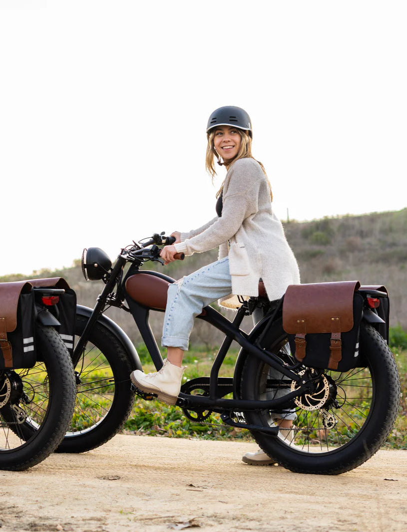 Two people standing next to vintage-style motorcycles on a dirt road with a scenic background.