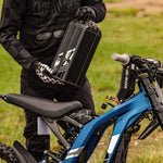 Person holding a surron 2025 lightbee x  black battery pack next to a blue electric bike in an outdoor setting
