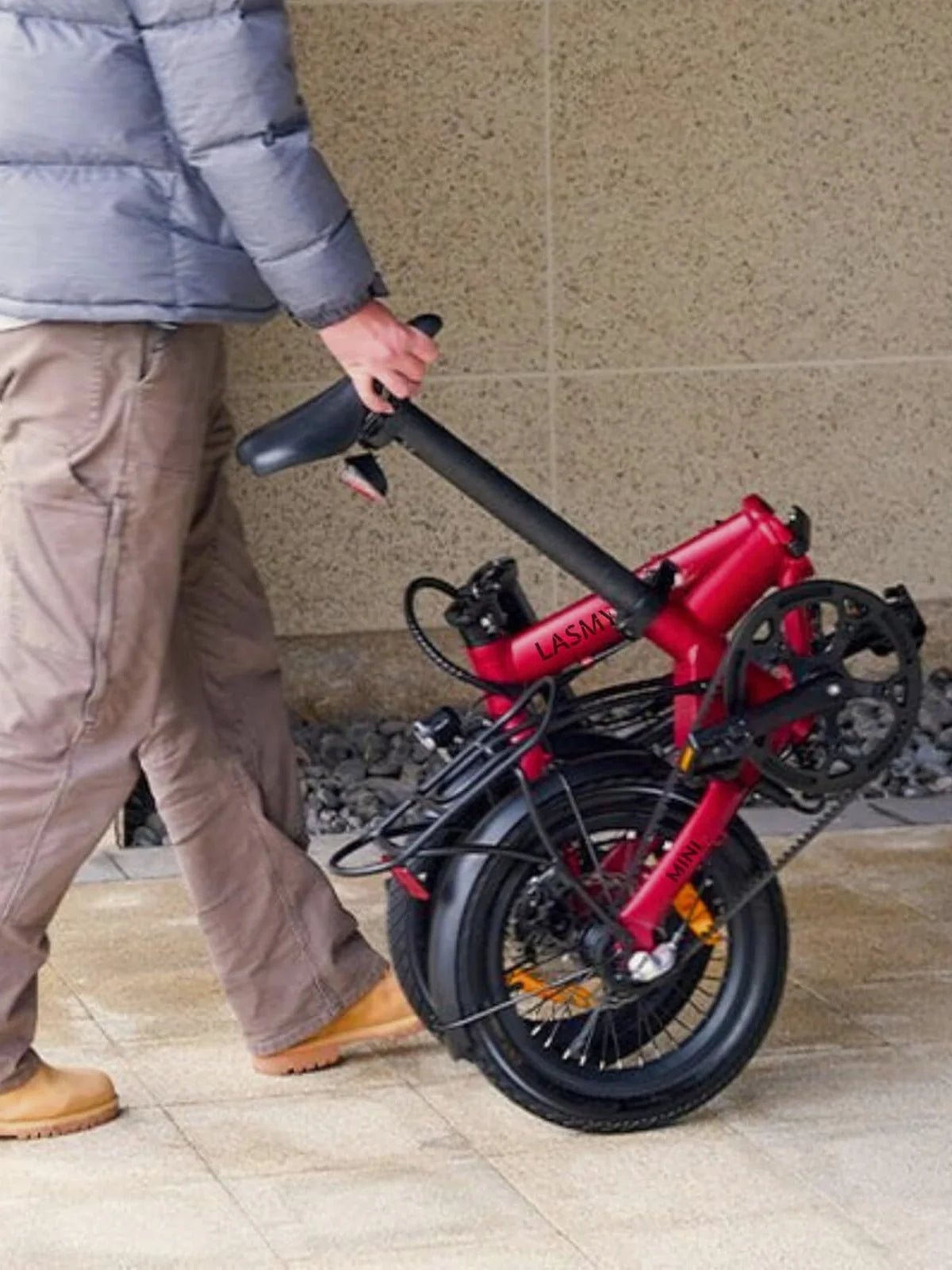 Person holding a red lasmy folding electric bike against a stone wall.
