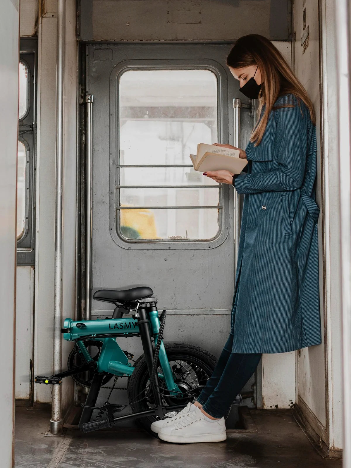 Woman in a blue coat reading a book on a train with a lasmy folding electric bike beside her.