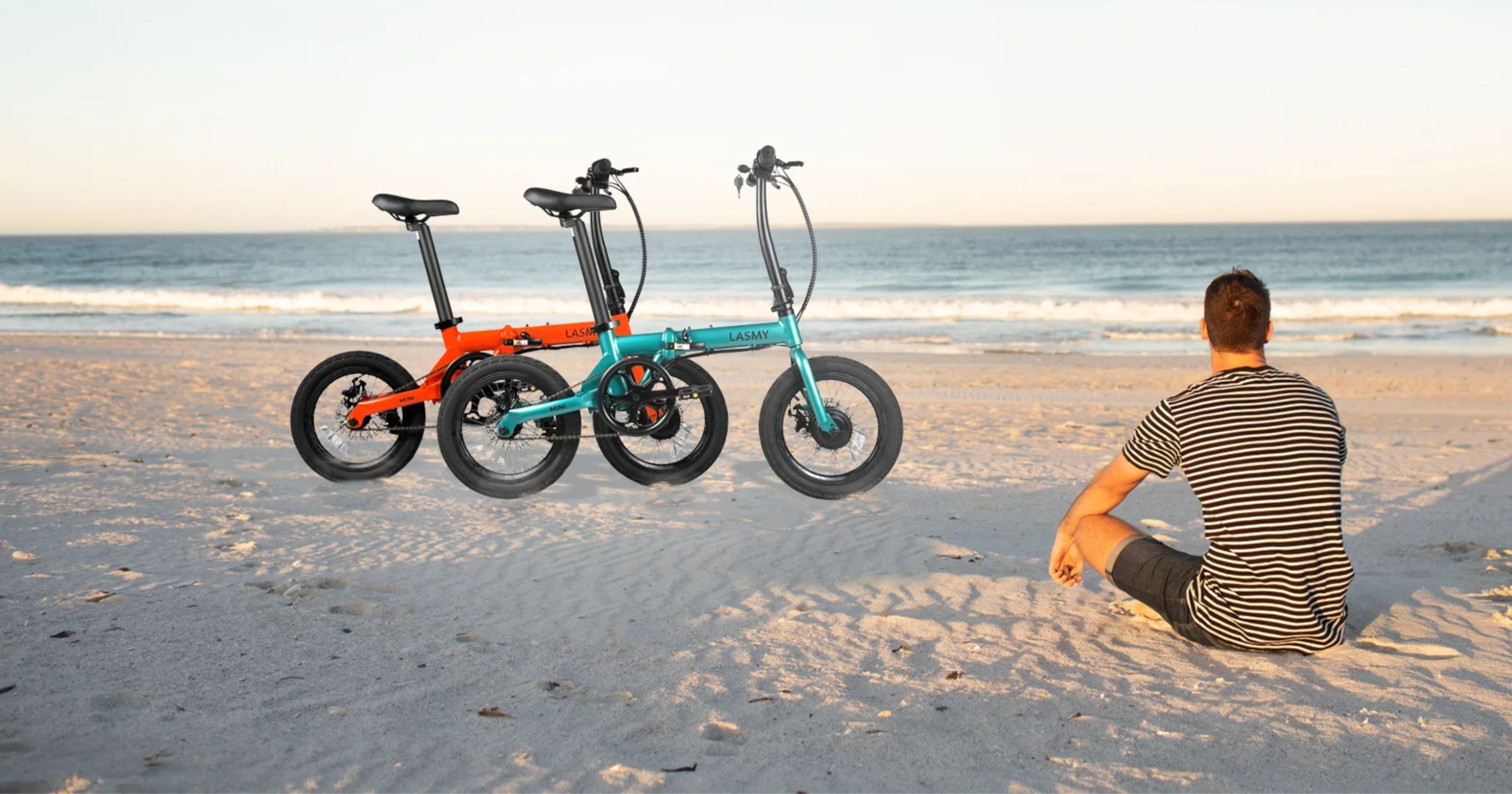 Two electric bikes on a sandy beach with a person sitting nearby.